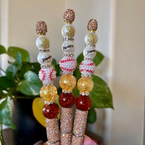 May include: Three decorative pens featuring a baseball theme. Each pen has a rhinestone-covered barrel, topped with beads in white, gold, and red, and a baseball-shaped bead. The pens also feature the San Diego Padres logo.