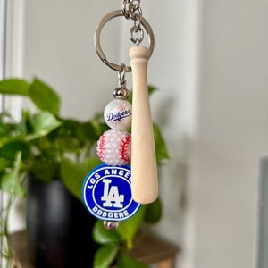 May include: A baseball-themed keychain featuring a wooden baseball bat charm, a white baseball bead, a pink beaded section, and a blue and white Los Angeles Dodgers logo charm. The keychain is attached to a silver keyring.