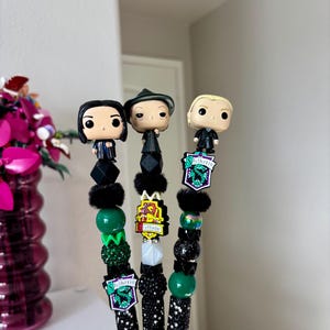 Peut inclure: Trois stylos décoratifs avec des personnages de Harry Potter. Chaque stylo a un corps noir recouvert de strass, orné de perles, de breloques et d'une figurine Funko Pop. Les stylos incluent les emblèmes des maisons Serpentard et Gryffondor.