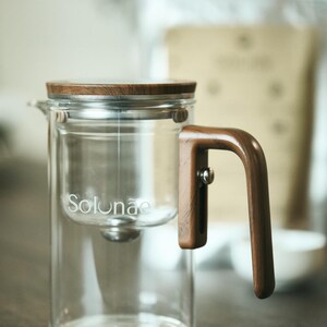 May include: A clear glass tea infuser with a wooden lid and handle. The infuser has a cylindrical shape with a built-in filter and the word "Solunae" printed on the side. The handle is made of dark wood. The tea infuser is designed for brewing loose leaf tea.