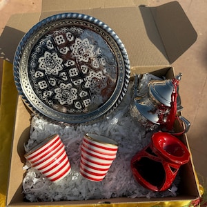 Pode incluir: Um conjunto de presente decorativo com uma bandeja prateada com um design floral ornamentado, um serviço de chá prateado, duas xícaras listradas em vermelho e branco e um queimador de incenso de cerâmica vermelha. Os itens estão dispostos em uma caixa de papelão.