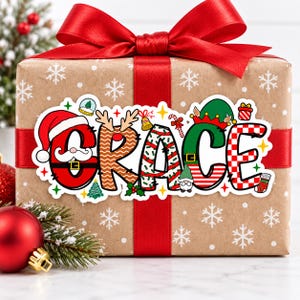 May include: A rectangular gift box wrapped in brown paper with white snowflakes, tied with a red ribbon and bow. A large sticker displays the name GRACE in colorful, festive holiday-themed letters featuring Santa, reindeer, elf, and candy cane motifs.