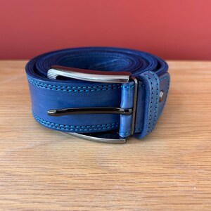 Puede incluir: Un cintur&oacute;n de cuero azul enrollado con una hebilla plateada. El cintur&oacute;n tiene costuras azul claro y una hebilla azul oscuro. El cintur&oacute;n est&aacute; sobre una superficie de madera con un fondo rojo.