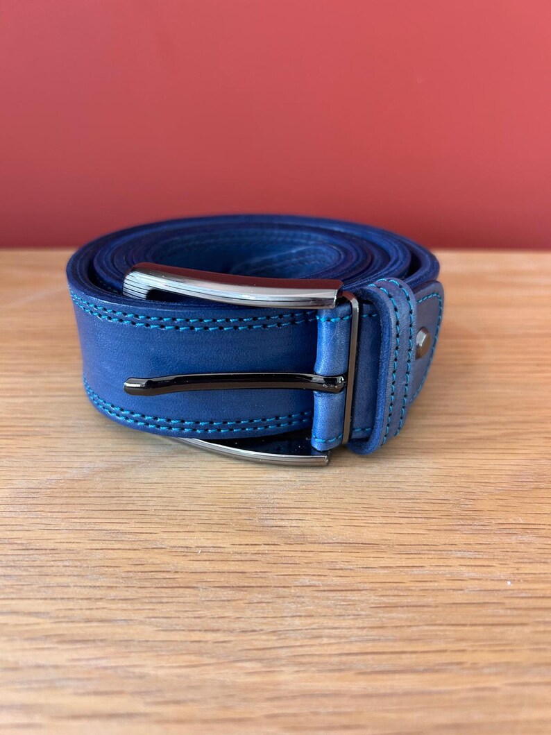 Puede incluir: Un cintur&oacute;n de cuero azul enrollado con una hebilla plateada. El cintur&oacute;n tiene costuras azul claro y una hebilla azul oscuro. El cintur&oacute;n est&aacute; sobre una superficie de madera con un fondo rojo.