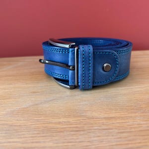 Puede incluir: Un cintur&oacute;n de cuero azul enrollado con una hebilla de color plateado y un remache a juego. El cintur&oacute;n tiene costuras azules en los bordes. El fondo es de color rojo liso.