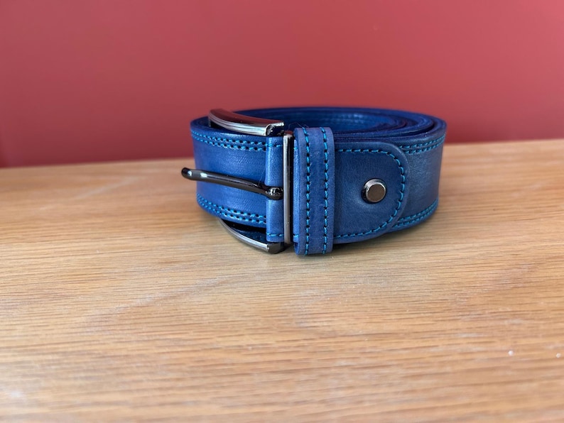 Puede incluir: Un cintur&oacute;n de cuero azul enrollado con una hebilla de color plateado y un remache a juego. El cintur&oacute;n tiene costuras azules en los bordes. El fondo es de color rojo liso.