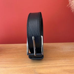 Puede incluir: Un cintur&oacute;n de cuero negro con hebilla plateada. El cintur&oacute;n est&aacute; enrollado y se muestra sobre una superficie de madera, con un fondo rojo. El cintur&oacute;n parece nuevo y sin usar.