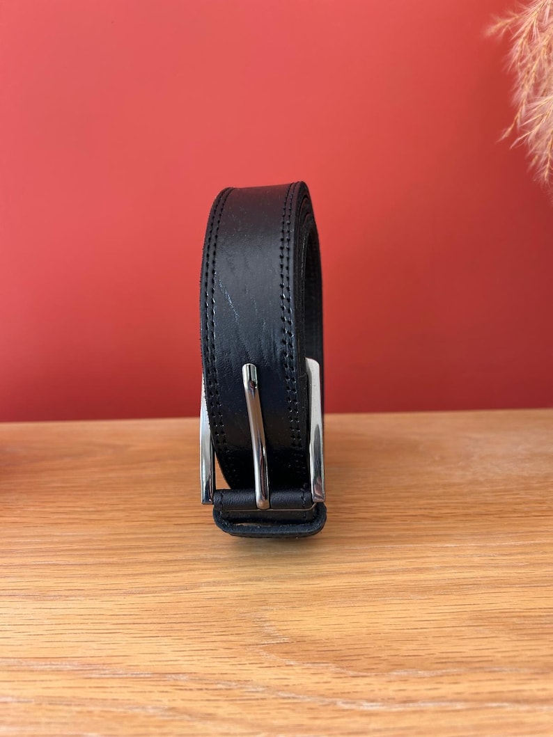 Puede incluir: Un cintur&oacute;n de cuero negro con hebilla plateada. El cintur&oacute;n est&aacute; enrollado y se muestra sobre una superficie de madera, con un fondo rojo. El cintur&oacute;n parece nuevo y sin usar.