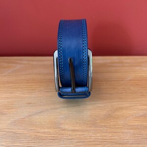 Puede incluir: Un cintur&oacute;n de cuero azul con una hebilla plateada. El cintur&oacute;n tiene un borde cosido y se muestra sobre una superficie de madera. El fondo es de color rojo liso.