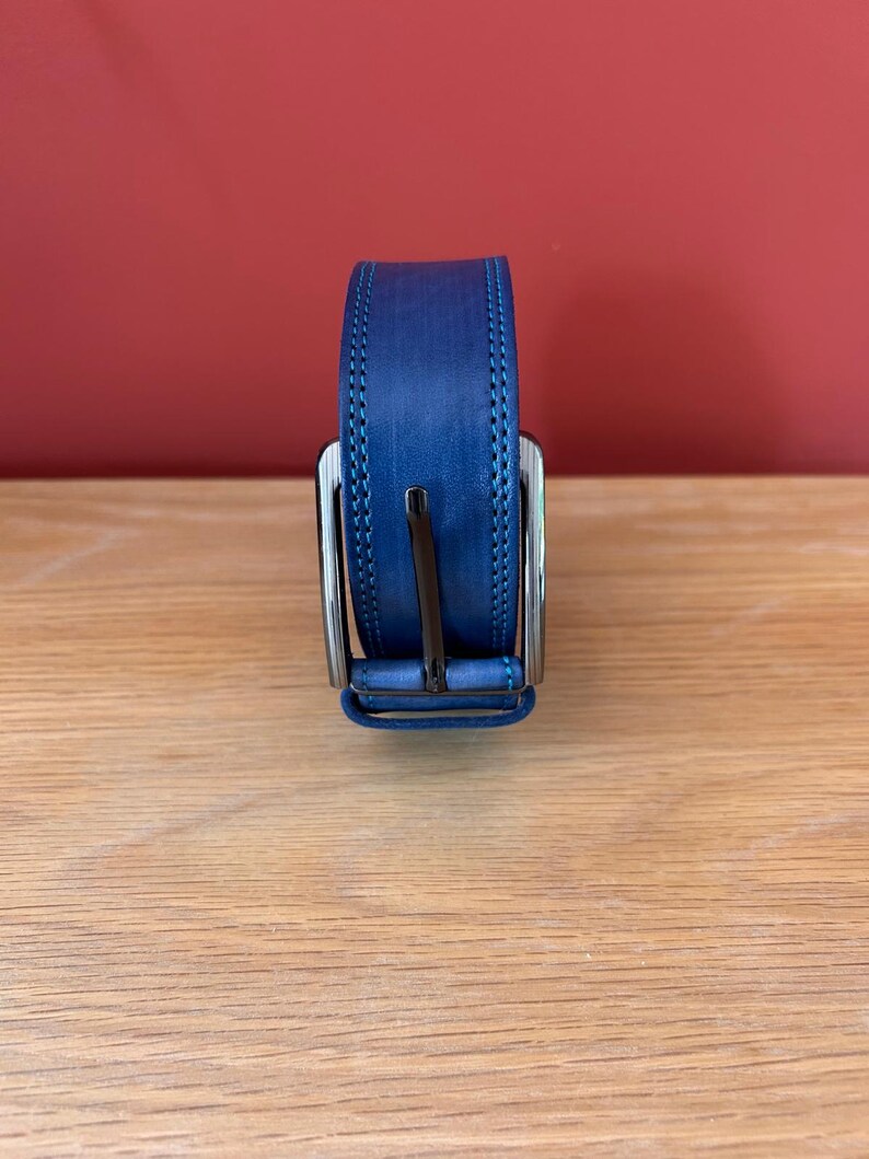 Puede incluir: Un cintur&oacute;n de cuero azul con una hebilla plateada. El cintur&oacute;n tiene un borde cosido y se muestra sobre una superficie de madera. El fondo es de color rojo liso.