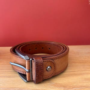 Puede incluir: Un cintur&oacute;n de cuero marr&oacute;n claro enrollado, con una hebilla plateada. El cintur&oacute;n est&aacute; sobre una superficie de madera con un fondo rojo. El cintur&oacute;n tiene un dise&ntilde;o cl&aacute;sico con costuras visibles y un remache met&aacute;lico.