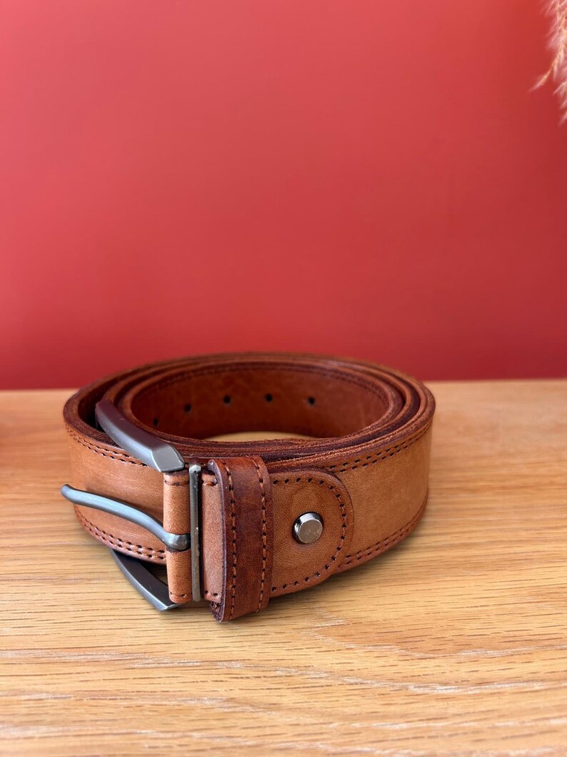 Puede incluir: Un cintur&oacute;n de cuero marr&oacute;n claro enrollado, con una hebilla plateada. El cintur&oacute;n est&aacute; sobre una superficie de madera con un fondo rojo. El cintur&oacute;n tiene un dise&ntilde;o cl&aacute;sico con costuras visibles y un remache met&aacute;lico.