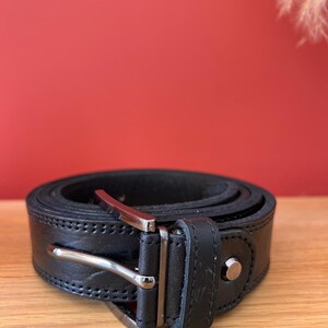 Puede incluir: Un cintur&oacute;n de cuero negro enrollado con una hebilla plateada. El cintur&oacute;n est&aacute; cosido a lo largo de los bordes y tiene un solo remache plateado. El fondo es de color rojo.