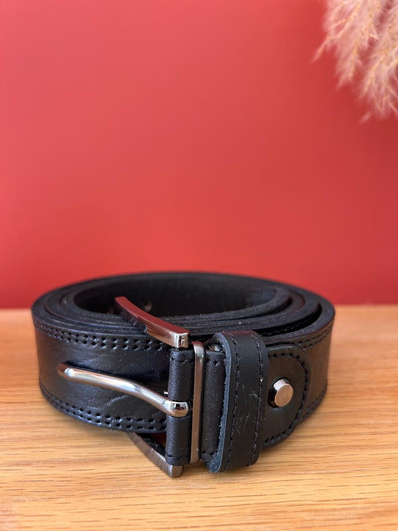 Puede incluir: Un cintur&oacute;n de cuero negro enrollado con una hebilla plateada. El cintur&oacute;n est&aacute; cosido a lo largo de los bordes y tiene un solo remache plateado. El fondo es de color rojo.
