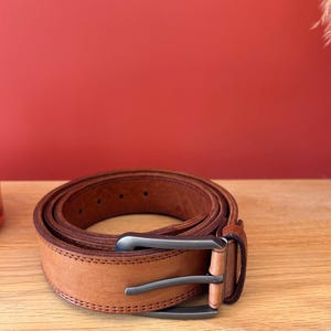 Puede incluir: Un cintur&oacute;n de cuero marr&oacute;n claro enrollado, con una hebilla plateada. El cintur&oacute;n est&aacute; sobre una superficie de madera con un fondo rojo. El cintur&oacute;n tiene un dise&ntilde;o cosido en los bordes.