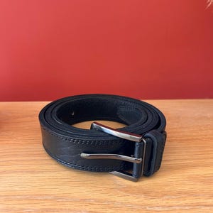 Puede incluir: Un cintur&oacute;n de cuero negro con una hebilla rectangular plateada. El cintur&oacute;n est&aacute; enrollado sobre una superficie de madera, mostrando su textura y costuras. El fondo es de color rojo liso.