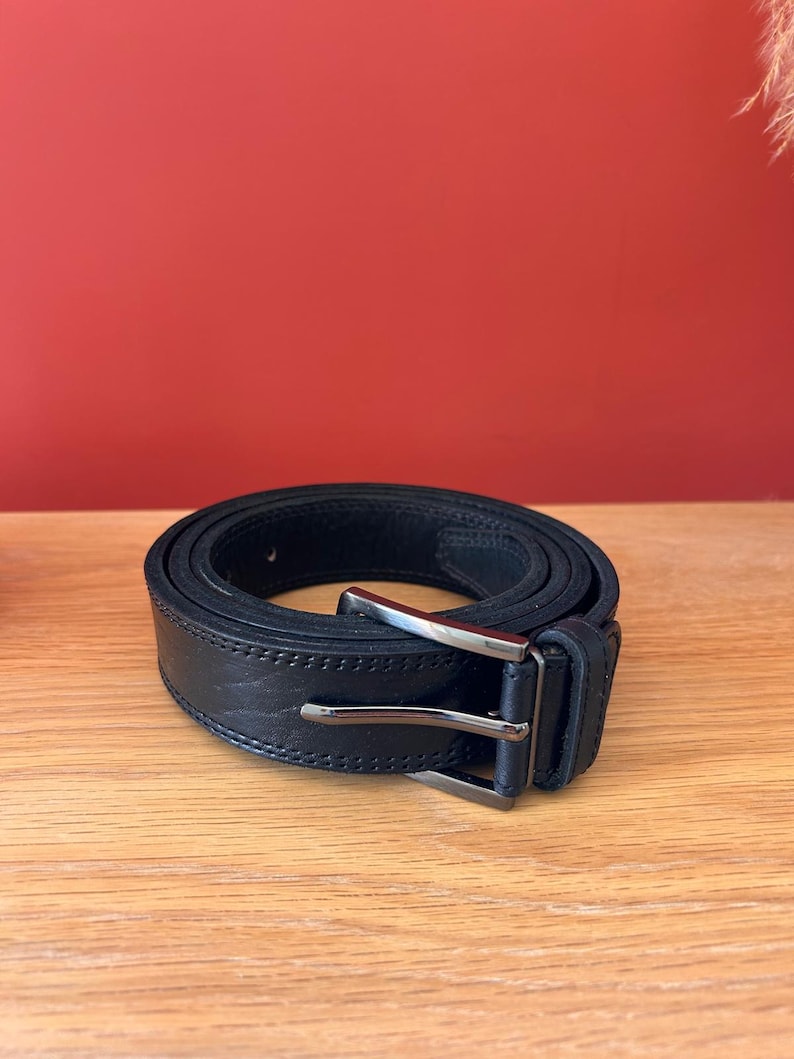 Puede incluir: Un cintur&oacute;n de cuero negro con una hebilla rectangular plateada. El cintur&oacute;n est&aacute; enrollado sobre una superficie de madera, mostrando su textura y costuras. El fondo es de color rojo liso.