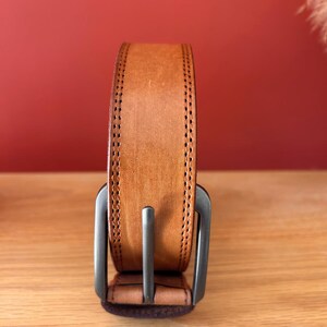 Puede incluir: Un cintur&oacute;n de cuero marr&oacute;n claro con una hebilla plateada. El cintur&oacute;n tiene un borde cosido y se muestra verticalmente sobre un fondo rojo. Un accesorio cl&aacute;sico.