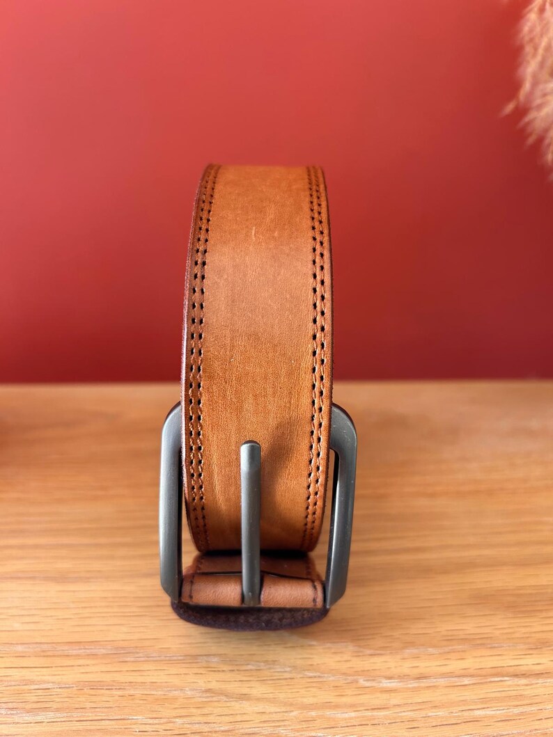 Puede incluir: Un cintur&oacute;n de cuero marr&oacute;n claro con una hebilla plateada. El cintur&oacute;n tiene un borde cosido y se muestra verticalmente sobre un fondo rojo. Un accesorio cl&aacute;sico.