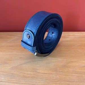 Puede incluir: Un cintur&oacute;n de cuero azul marino enrollado, con una hebilla plateada. El cintur&oacute;n tiene un borde cosido y un peque&ntilde;o tachuela plateada en la trabilla. El fondo es de color rojo.