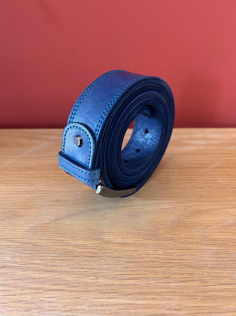 Puede incluir: Un cintur&oacute;n de cuero azul marino enrollado, con una hebilla plateada. El cintur&oacute;n tiene un borde cosido y un peque&ntilde;o tachuela plateada en la trabilla. El fondo es de color rojo.