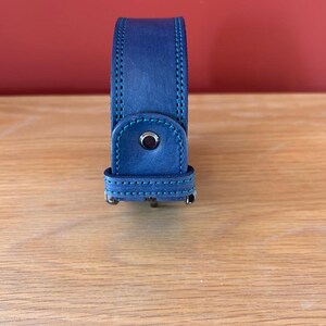 Puede incluir: Una correa de reloj de cuero azul con costuras turquesas. La correa tiene un cierre de hebilla y un detalle de bot&oacute;n oscuro. El fondo es una pared roja lisa y una superficie de madera.