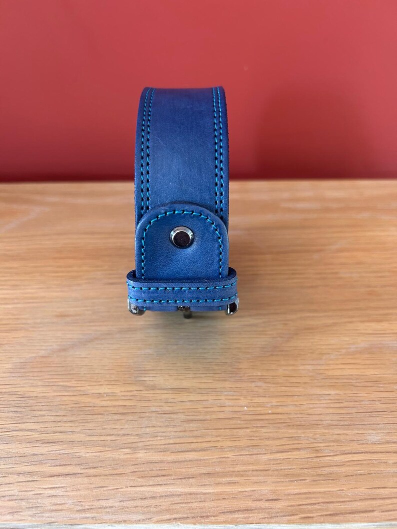 Puede incluir: Una correa de reloj de cuero azul con costuras turquesas. La correa tiene un cierre de hebilla y un detalle de bot&oacute;n oscuro. El fondo es una pared roja lisa y una superficie de madera.