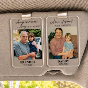 May include: Two rectangular clear acrylic car visor clips featuring printed photographs and text. The left clip shows a man and child with the text Always in my heart, Always on my journey, GRANDPA, 1939-2026. The right clip shows a man and child with the text A piece of my heart RIDES IN HEAVEN, DADDY, 1976-2026.