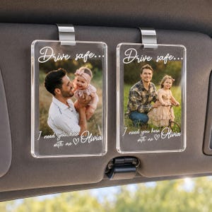 May include: Two rectangular clear acrylic car visor clips featuring custom photo prints of a person with a child. The text "Drive safe... I need you here with me Olivia" is printed in white script on the surface of each transparent plaque.