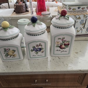 May include: Three white ceramic jars with fruit-themed lids and labels. The lids are topped with a lemon, a blueberry, and a red apple. The labels feature illustrations of lemons, plums, and apples, with botanical names.