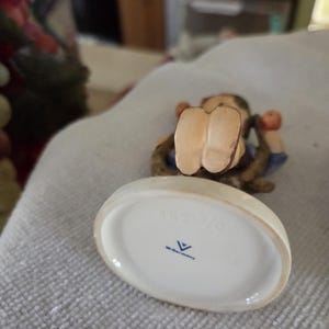 May include: A ceramic figurine of a child lying down, with visible feet and arms. The figurine is wearing blue clothing and has brown hair. The base is white with the text "142 3/0" and a logo.