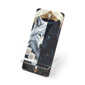 May include: A phone stand featuring a split design with a white wolf on the left and a black wolf on the right. The wolves have blue eyes. White roses are on the left, and black roses are on the right. The stand has a clear base.