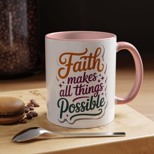 Könnte beinhalten: Weißer Keramikbecher mit rosa Innenraum und Henkel. Der Becher trägt den Text "Faith makes all things Possible" in orange, lila und grün. Ein Löffel und ein Gebäck liegen auf einer Holzoberfläche.