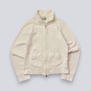 以下が含まれることがあります： クリーム色のジップアップジャケット。ハイカラーと2つのフロントポケットが付いています。胸元には小さなナイキのロゴがあり、フルレングスのジッパーが付いています。生地は柔らかいリブ素材のようです。