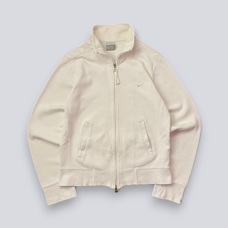 以下が含まれることがあります： クリーム色のジップアップジャケット。ハイカラーと2つのフロントポケットが付いています。胸元には小さなナイキのロゴがあり、フルレングスのジッパーが付いています。生地は柔らかいリブ素材のようです。
