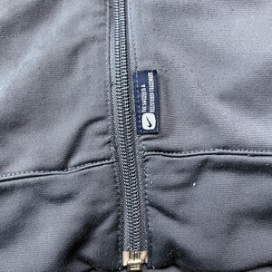 May include: Close-up of a dark gray athletic jacket with a full-length zipper. A small tag with the Nike logo and the text "THE SWOOSH IS A REGISTERED TRADEMARK" is sewn into the jacket.