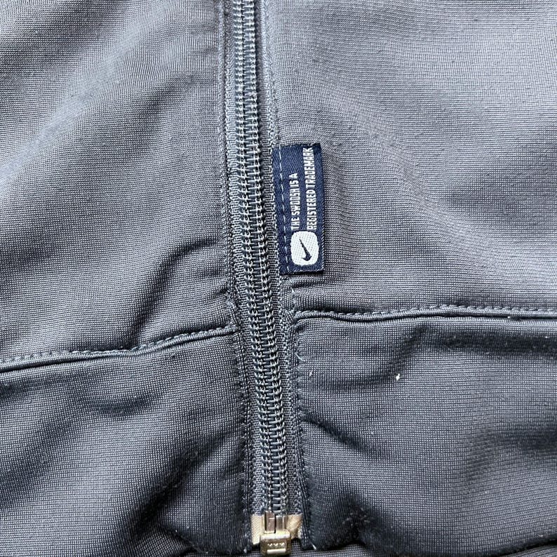 May include: Close-up of a dark gray athletic jacket with a full-length zipper. A small tag with the Nike logo and the text "THE SWOOSH IS A REGISTERED TRADEMARK" is sewn into the jacket.