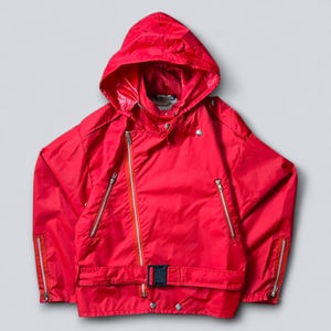 Puede incluir: Chaqueta roja brillante con capucha, múltiples cremalleras plateadas, cinturón con hebilla negra y cierres a presión plateados. La chaqueta tiene capucha y bolsillos con cremallera. El tejido es brillante y ligero.