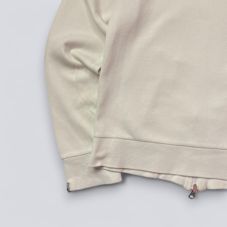 以下が含まれることがあります： 薄いベージュ色のジップアップスウェットシャツ。 スウェットシャツは、リブ編みの裾と袖口、金属製のジッパーが付いています。 生地は柔らかいニット素材のようです。 袖は画像左側に表示されています。