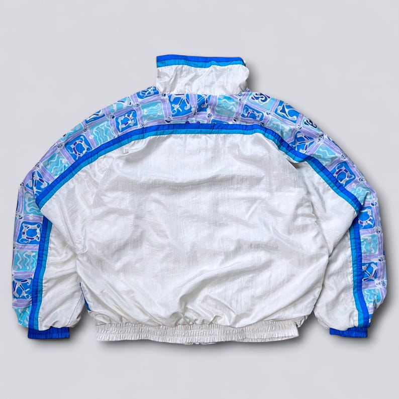May include: White vintage jacket with a high collar and elastic cuffs. The jacket features a geometric pattern in shades of blue and purple along the shoulders and sleeves. A blue stripe runs across the back. The jacket has a full zipper and a drawstring hem.