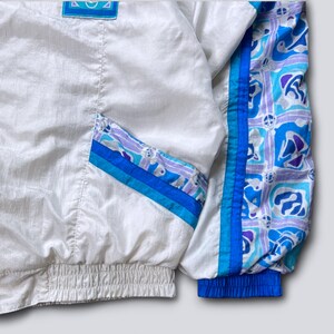 May include: White vintage windbreaker jacket with blue and purple patterned accents. Features a blue elastic cuff and waist. The jacket has a front pocket with blue and purple stripes.
