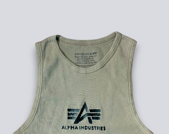 May include: A sage green ribbed tank top featuring a dark navy blue Alpha Industries logo printed on the chest. The interior neckline displays printed manufacturer text including Alpha Industries Inc, Knoxville, Tennessee, USA.