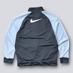 May include: A dark gray track jacket with light blue sleeves. The jacket features a white Nike logo on the back and a full-length zipper. The cuffs and collar are dark gray.