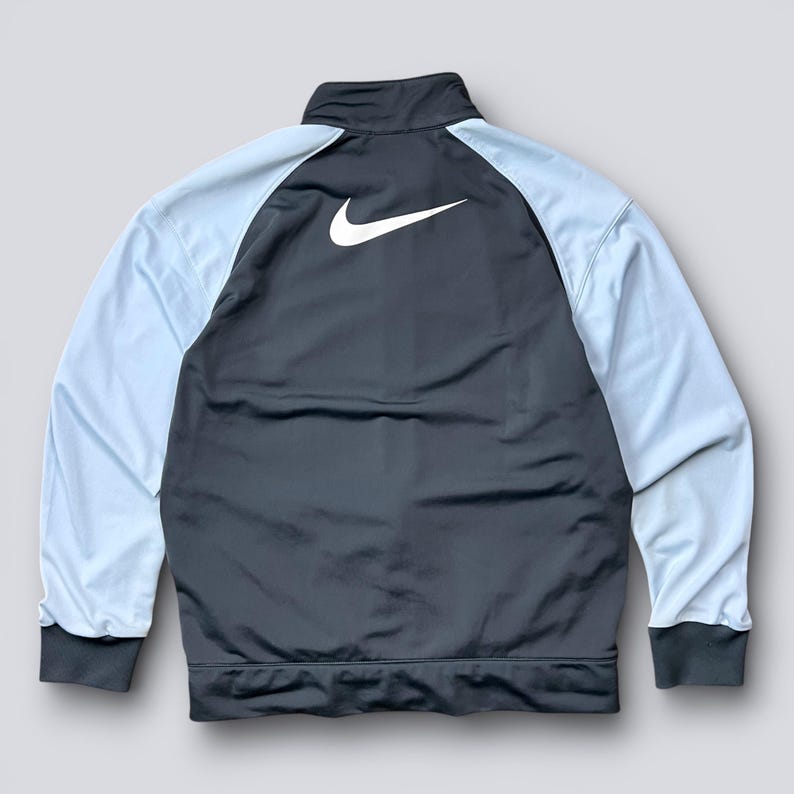 May include: A dark gray track jacket with light blue sleeves. The jacket features a white Nike logo on the back and a full-length zipper. The cuffs and collar are dark gray.