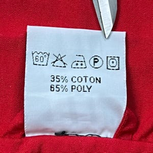 May include: A close-up of a white clothing label with black symbols and text. The text reads "35% COTON" and "65% POLY". The label is on a red fabric, and a pair of scissors is partially visible.