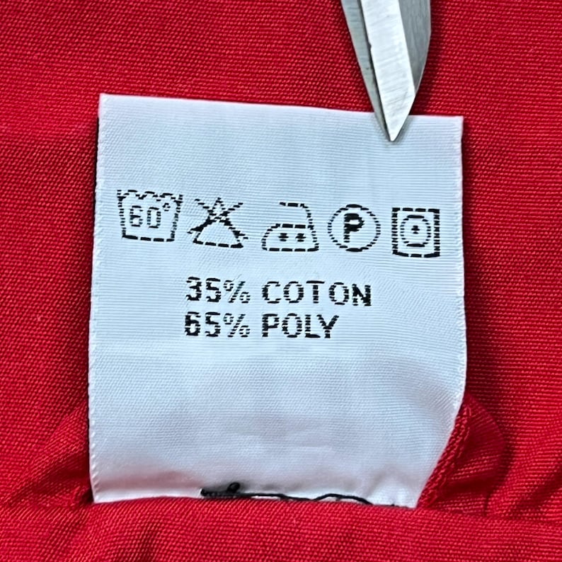 May include: A close-up of a white clothing label with black symbols and text. The text reads "35% COTON" and "65% POLY". The label is on a red fabric, and a pair of scissors is partially visible.
