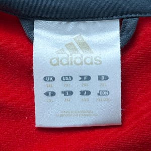 May include: Close-up of a white Adidas clothing tag with the Adidas logo and size information. The tag is against a red fabric background. The tag includes size designations for UK, USA, F, D, E, I, J, and CDN.