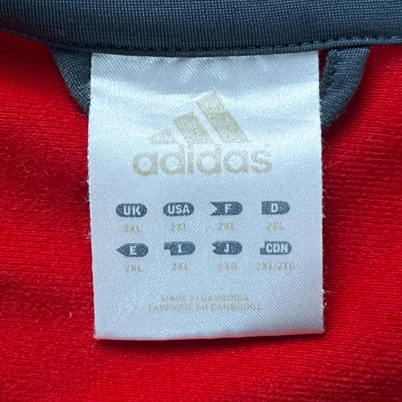 May include: Close-up of a white Adidas clothing tag with the Adidas logo and size information. The tag is against a red fabric background. The tag includes size designations for UK, USA, F, D, E, I, J, and CDN.