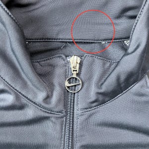 May include: Close-up of a black athletic jacket with a zipper and a Nike logo on the zipper pull. The jacket has a high collar and a shiny, smooth texture. The stitching is visible along the seams.