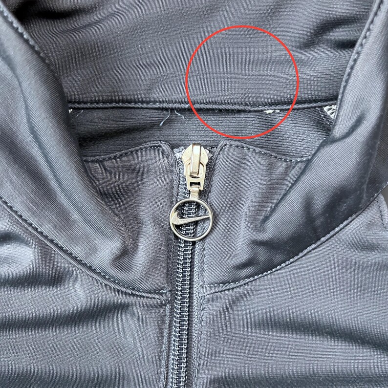 May include: Close-up of a black athletic jacket with a zipper and a Nike logo on the zipper pull. The jacket has a high collar and a shiny, smooth texture. The stitching is visible along the seams.
